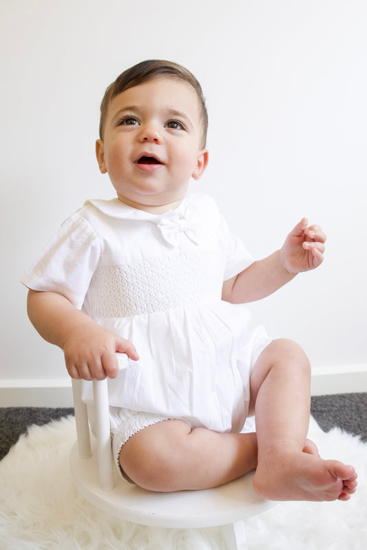 Baby in a white outfit sitting on a chair with a white background